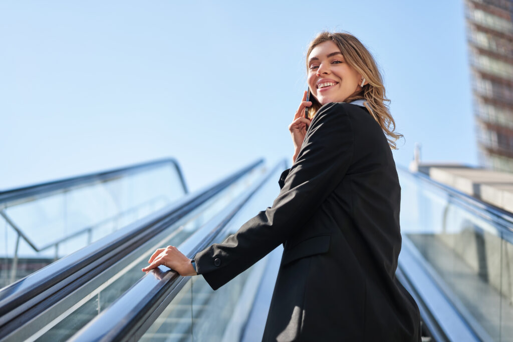 portrait of businesswoman in black suit, going up on escalator, talking on mobile phone. saleswoman walking in city, having a call