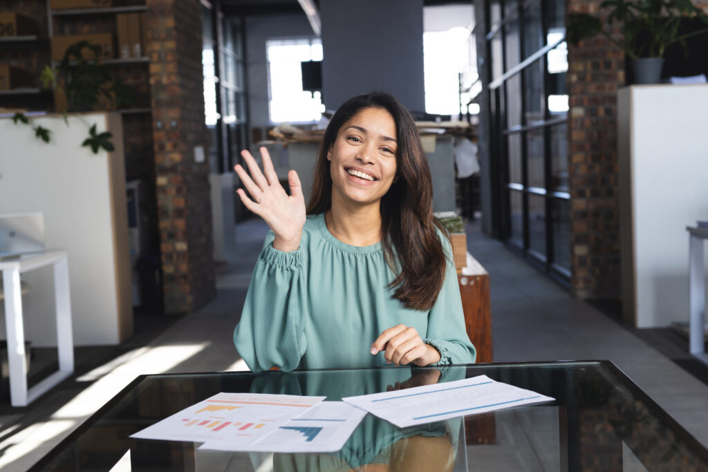 portrait of happy biracial casual businesswoman having video call and waving hand in office. business, work, technology and communication, unaltered.